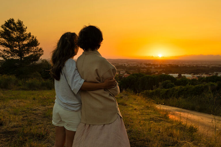 A woman stands with her aging mother, concerned about sundowning triggers as the sun starts to set.