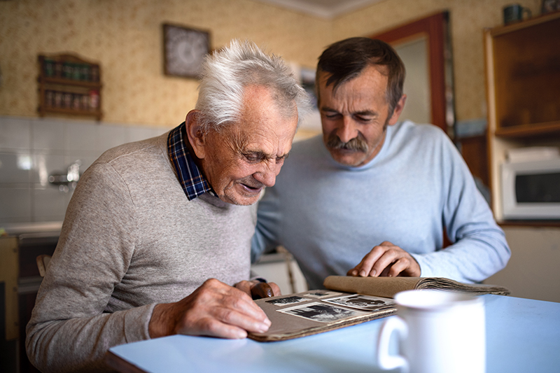A senior man smiles while looking at a photo album with his son who is interested in how dementia and sports memories can reignite emotional memory in dementia.
